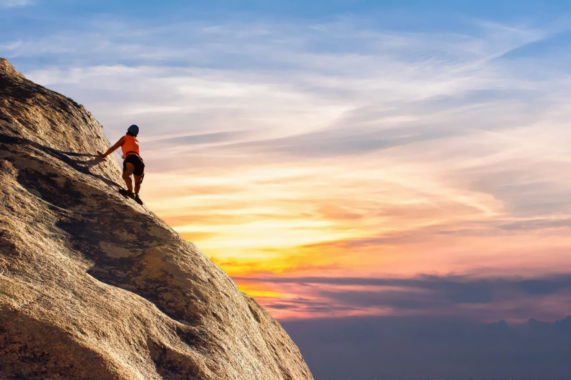Ein mutiger Kletterer erklimmt eine steile Felswand, sein Blick fest auf den Gipfel gerichtet, während der Himmel im Hintergrund in atemberaubenden Orange- und Blautönen leuchtet. Diese Szene verkörpert die Reise der Selbstfindung, symbolisiert durch den Mut, sich Herausforderungen zu stellen und über persönliche Grenzen hinauszugehen.