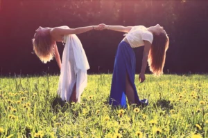 Zwei Frauen, verbunden durch das Halten der Hände, neigen sich rückwärts in einer tiefen Verbeugung in einem sonnendurchfluteten Feld, symbolisieren Achtsamkeit und Selbstfindung durch ihre harmonische Verbindung mit der Natur.