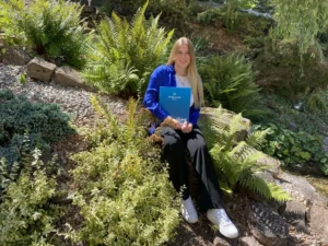 Eine junge Auszubildende zur Veranstaltungskauffrau sitzt lächelnd in einem grünen Garten, stolz ein Zertifikat der Akademie Heiligenfeld haltend, umgeben von einer Vielfalt an Pflanzen und einem Gefühl von Erfüllung und Neubeginn ausstrahlend.