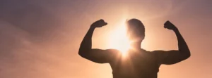 Die Silhouette eines muskulösen Mannes steht vor einem strahlenden Sonnenuntergang, die Arme in einer Pose der Stärke und des Triumphs erhoben. Diese kraftvolle Szene symbolisiert Resilienz und innere Stärke, Schlüsselelemente in der Psychotherapie, die Menschen dabei unterstützt, Herausforderungen zu überwinden und persönliches Wachstum zu fördern.