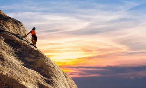 Ein mutiger Kletterer erklimmt eine steile Felswand, sein Blick fest auf den Gipfel gerichtet, während der Himmel im Hintergrund in atemberaubenden Orange- und Blautönen leuchtet. Diese Szene verkörpert die Reise der Selbstfindung, symbolisiert durch den Mut, sich Herausforderungen zu stellen und über persönliche Grenzen hinauszugehen.