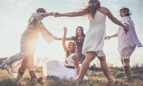 Eine Gruppe von Frauen genießt gemeinsam eine Auszeit in der Natur, umgeben von einem Feld bei Sonnenuntergang. Sie halten sich an den Händen, tanzen und lachen in einem Kreis, während eine von ihnen sich in der Mitte dreht. Die Szene strahlt Freude, Freiheit und Gemeinschaft aus, während jede Frau ihren eigenen Weg der Selbstfindung geht. Ihre Kleidung und Accessoires, von Blumenkränzen bis zu luftigen Kleidern, unterstreichen die ungezwungene und spirituelle Atmosphäre dieses besonderen Moments der Verbindung und des persönlichen Wachstums.