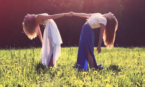 Zwei Frauen, verbunden durch das Halten der Hände, neigen sich rückwärts in einer tiefen Verbeugung in einem sonnendurchfluteten Feld, symbolisieren Achtsamkeit und Selbstfindung durch ihre harmonische Verbindung mit der Natur.