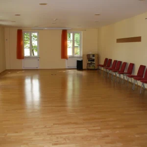 Ein leerer, heller Seminarraum in der Akademie Heiligenfeld mit glänzendem Holzboden und einer Reihe roter Stühle an der Seite. Die großen Fenster sind mit orangefarbenen Vorhängen versehen, die für eine warme und einladende Atmosphäre sorgen, ideal für Seminare und Workshops.