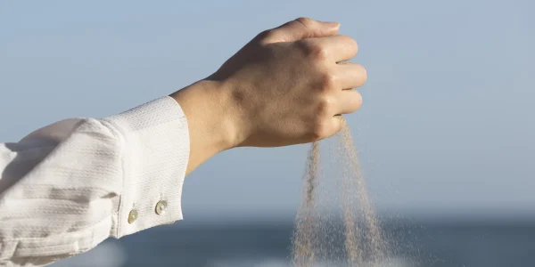 Eine Hand lässt feinen Sand behutsam durch die Finger rieseln, was die feine Wahrnehmung und Hochsensibilität symbolisiert, eingefangen vor dem unscharfen Hintergrund eines ruhigen Strandes.