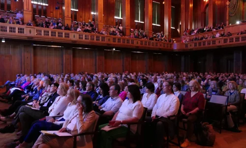 Ein aufmerksames Publikum sitzt in einem prachtvollen Saal, die Blicke gebannt auf die Bühne gerichtet, wo ein Vortrag im Rahmen eines Kongresses stattfindet. Die Teilnehmer verkörpern Offenheit und Bereitschaft zum Dialog und Wissensaustausch, eingetaucht in die inspirierende Atmosphäre des Kongresses.