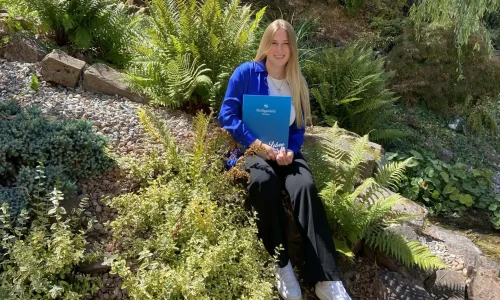 Eine junge Auszubildende zur Veranstaltungskauffrau sitzt lächelnd in einem grünen Garten, stolz ein Zertifikat der Akademie Heiligenfeld haltend, umgeben von einer Vielfalt an Pflanzen und einem Gefühl von Erfüllung und Neubeginn ausstrahlend.