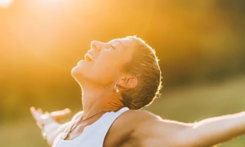 Ein Moment der Offenheit, eingefangen in einem Bild: Eine Person steht in der goldenen Stunde des Sonnenuntergangs, Arme weit geöffnet, das Gesicht zur Sonne erhoben, in einem Ausdruck purer Freude und Befreiung. Die Wärme des Lichts umhüllt die Szene, unterstreicht das Gefühl der Verbundenheit mit der Natur und der Welt. Ein Sinnbild für die Offenheit gegenüber den Geschenken des Lebens.