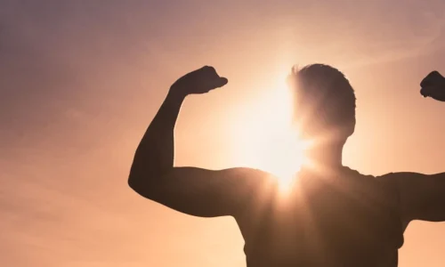 Die Silhouette eines muskulösen Mannes steht vor einem strahlenden Sonnenuntergang, die Arme in einer Pose der Stärke und des Triumphs erhoben. Diese kraftvolle Szene symbolisiert Resilienz und innere Stärke, Schlüsselelemente in der Psychotherapie, die Menschen dabei unterstützt, Herausforderungen zu überwinden und persönliches Wachstum zu fördern.