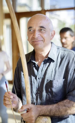 Ein Mann mit einem freundlichen Lächeln hält in einem hellen, offenen Raum mit großen Fenstern einen Stock und einen Schlegel, bereit, einen TaKeTiNa®-Rhythmus-Workshop zu leiten, während im Hintergrund Teilnehmende in Erwartung der nächsten Übung stehen.