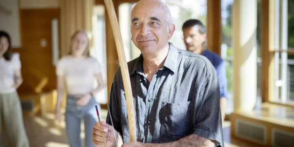 Ein Mann mit einem freundlichen Lächeln hält in einem hellen, offenen Raum mit großen Fenstern einen Stock und einen Schlegel, bereit, einen TaKeTiNa®-Rhythmus-Workshop zu leiten, während im Hintergrund Teilnehmende in Erwartung der nächsten Übung stehen.